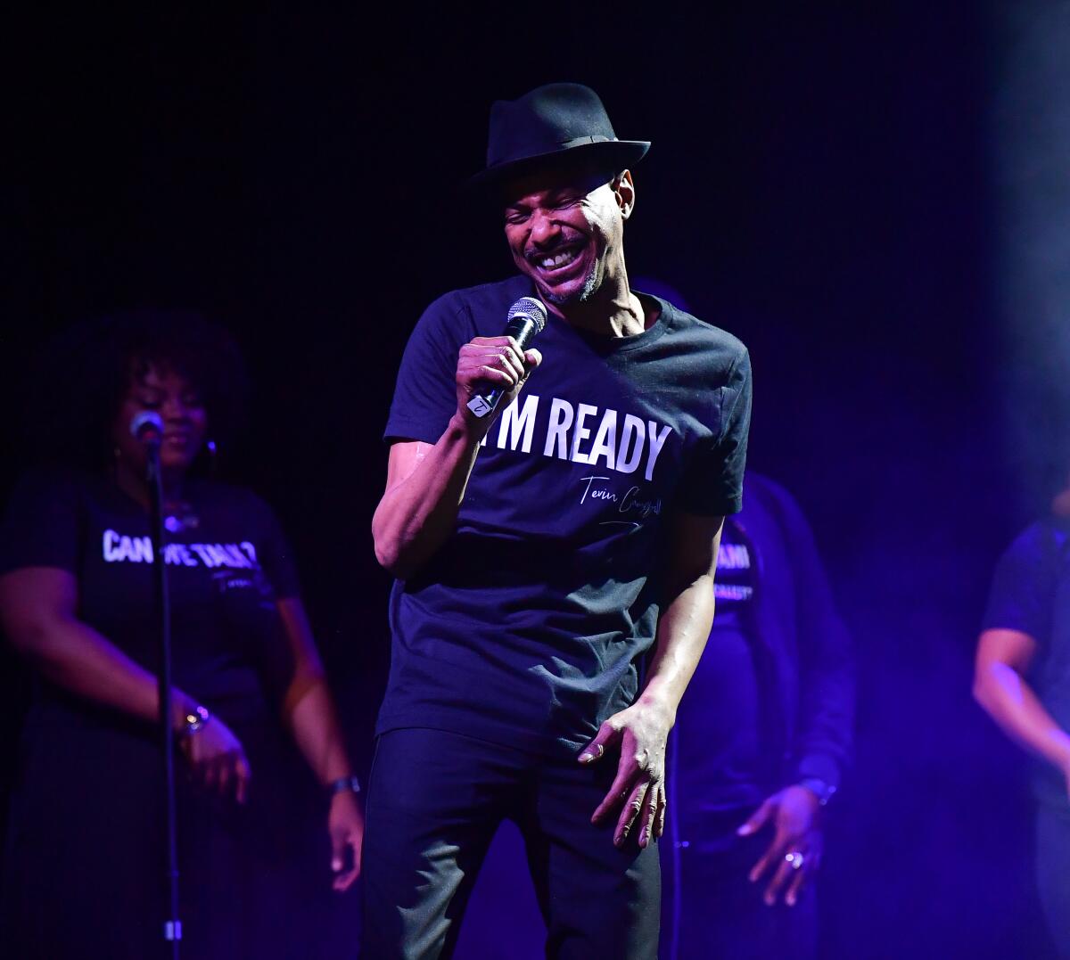 A man wearing all black and holding a microphone sings on a stage.