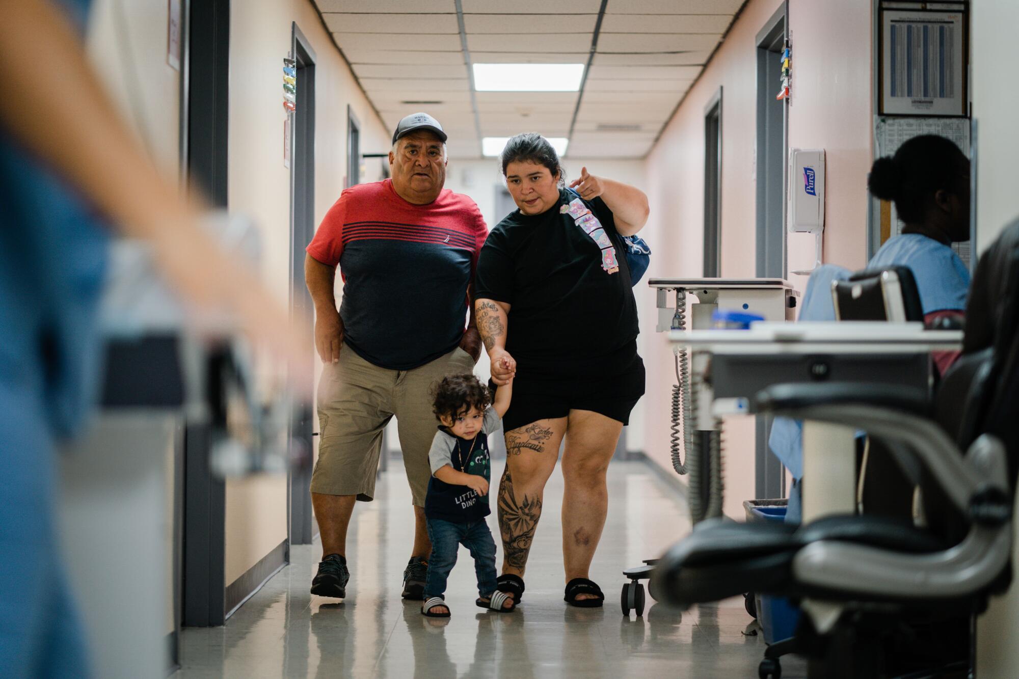 An older man and a young woman leading a toddler walk together down a hospital hallway. 
