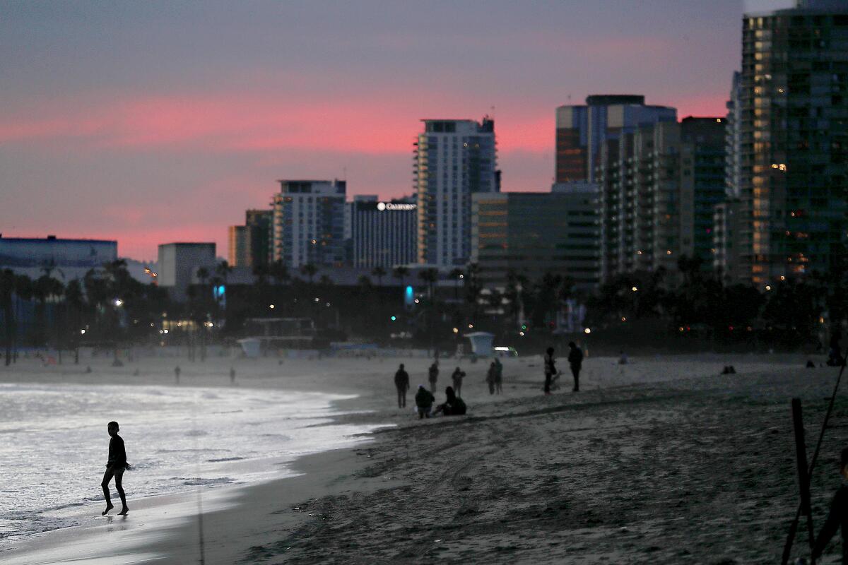 People are silhouetted at dusk on a beach with city buildings in the background.