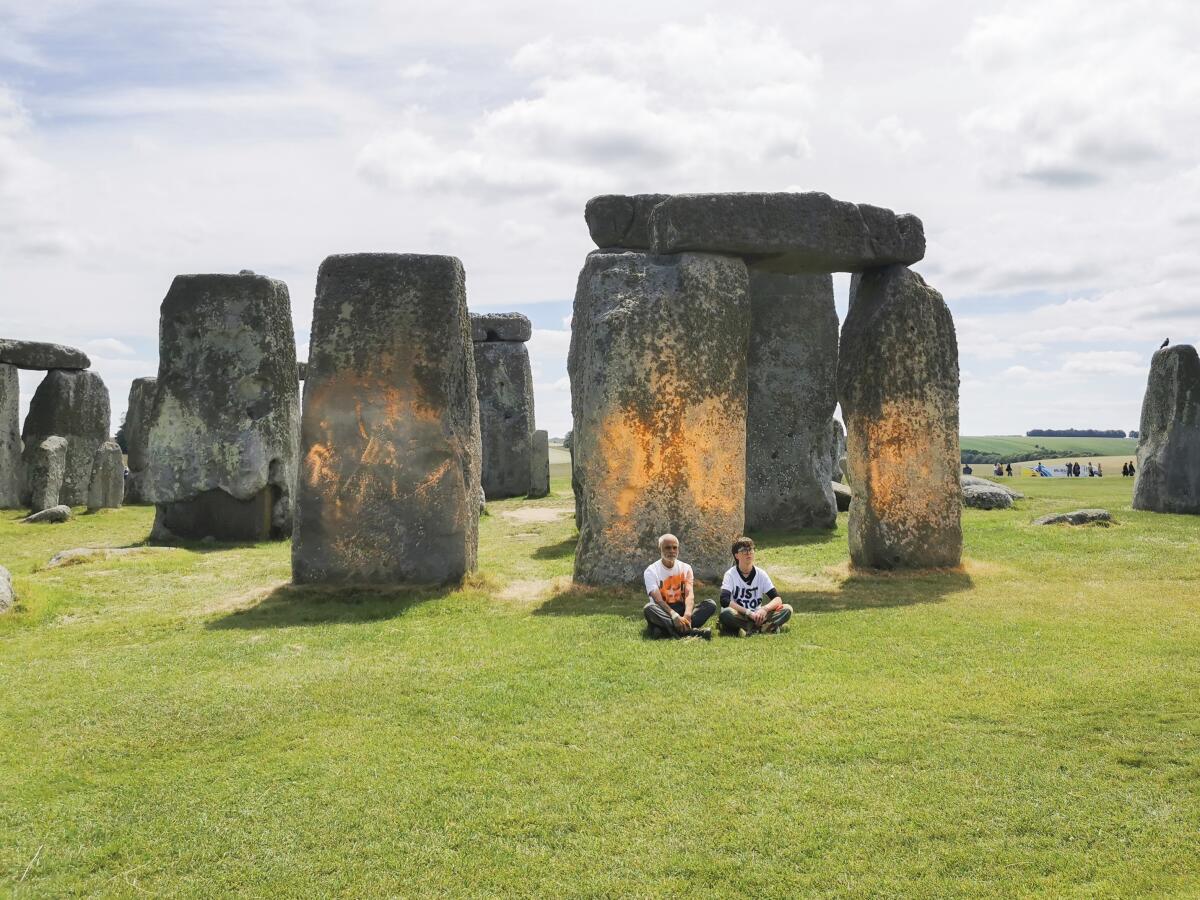 Two people sit cross-legged on the ground in front of orange-smudged  monoliths of Stonehenge