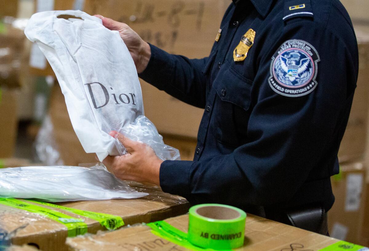 A U.S. Customs and Border Protection officer holds a counterfeit Dior shirt while he inspects boxes of products 