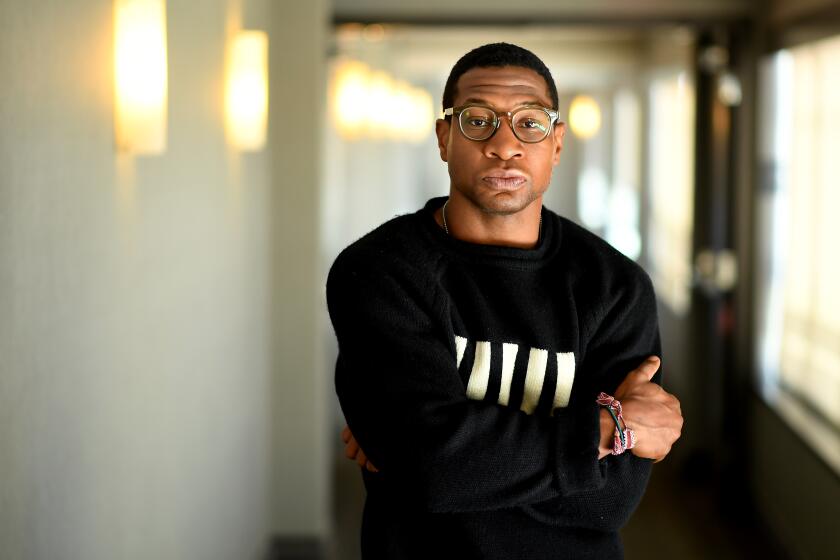 Jonathan Major in glasses and a a black sweater with thick white lines on it posing with his arms crossed