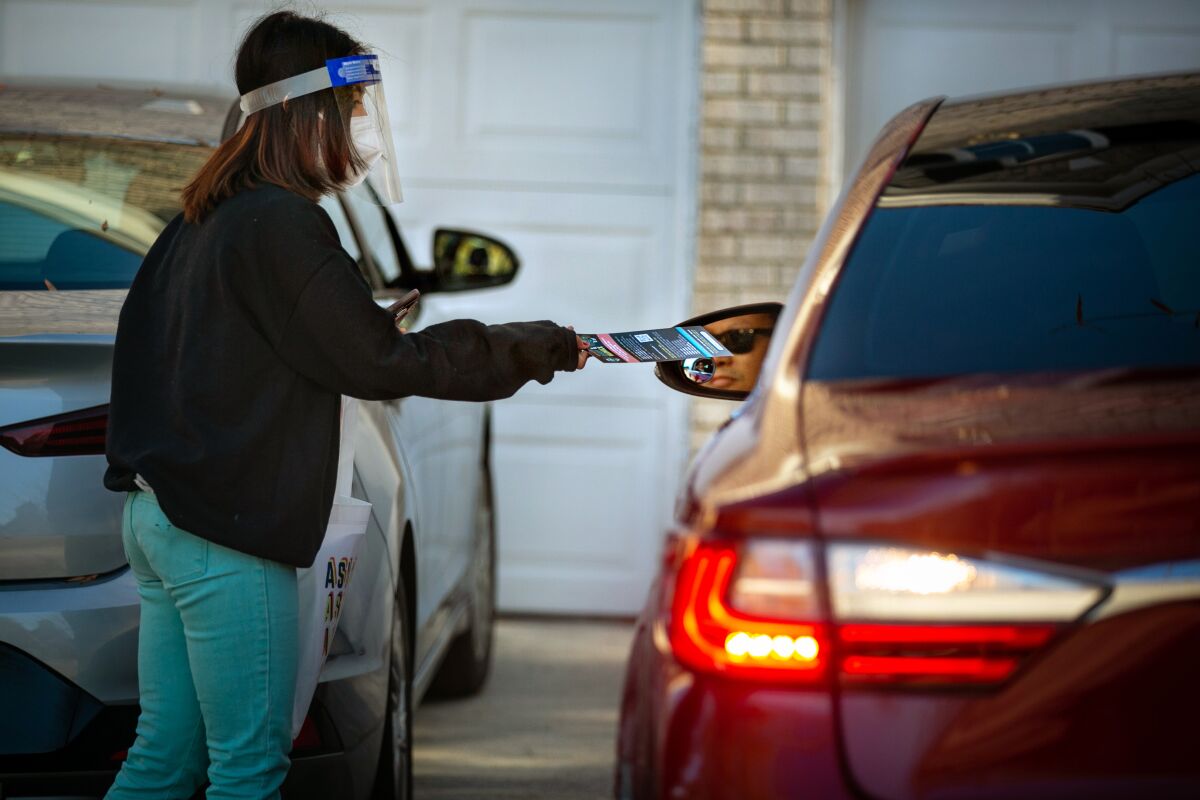 A woman in mask and face shield hands a pamphlet to a person in a car.