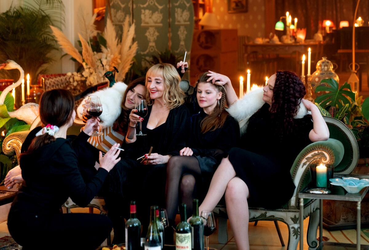Several women in fancy black dress sit on an ornate sofa while another woman sits nearby with a wine glass.