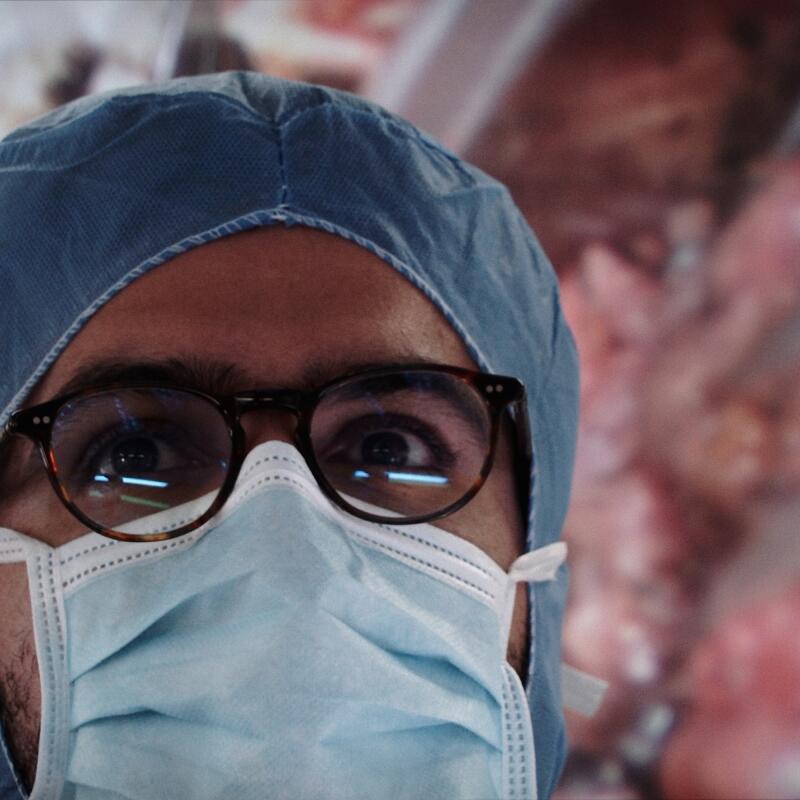 A surgeon's face, in hat, mask and glasses, with a view of the patient he's working on above him