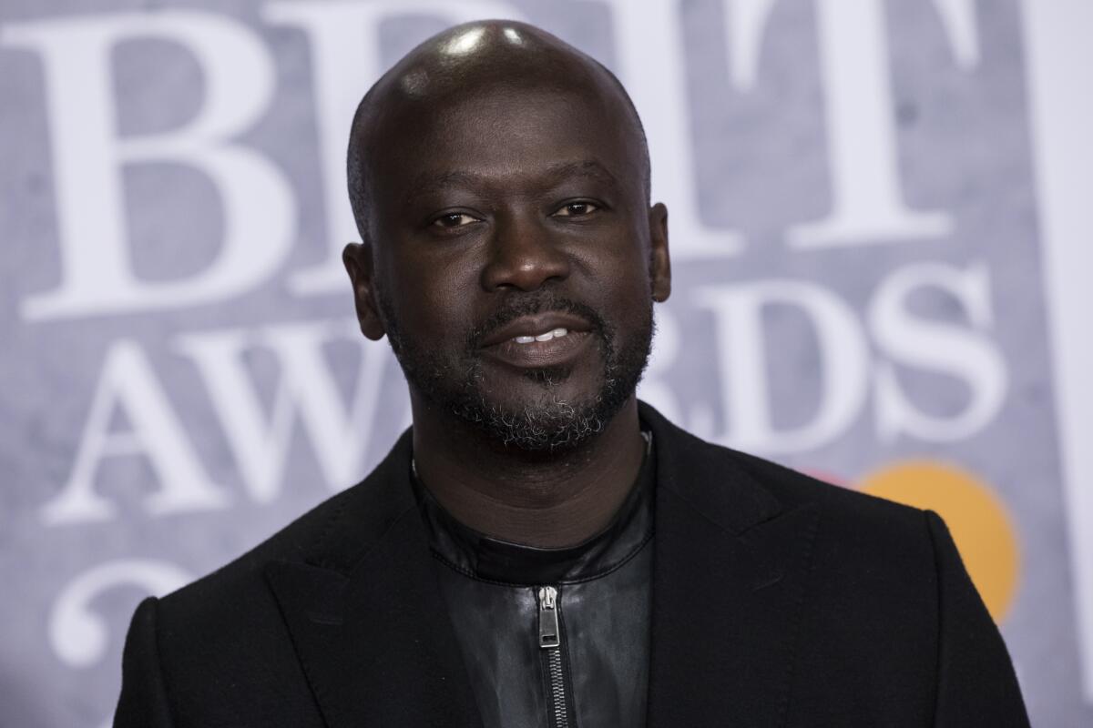 David Adjaye, wearing a dark suit, stands before a gray scrim at an awards ceremony.