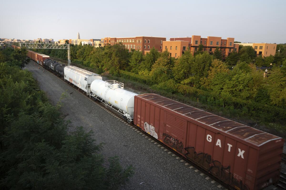 A freight train passes by trees to its right and a cluster of buildings.