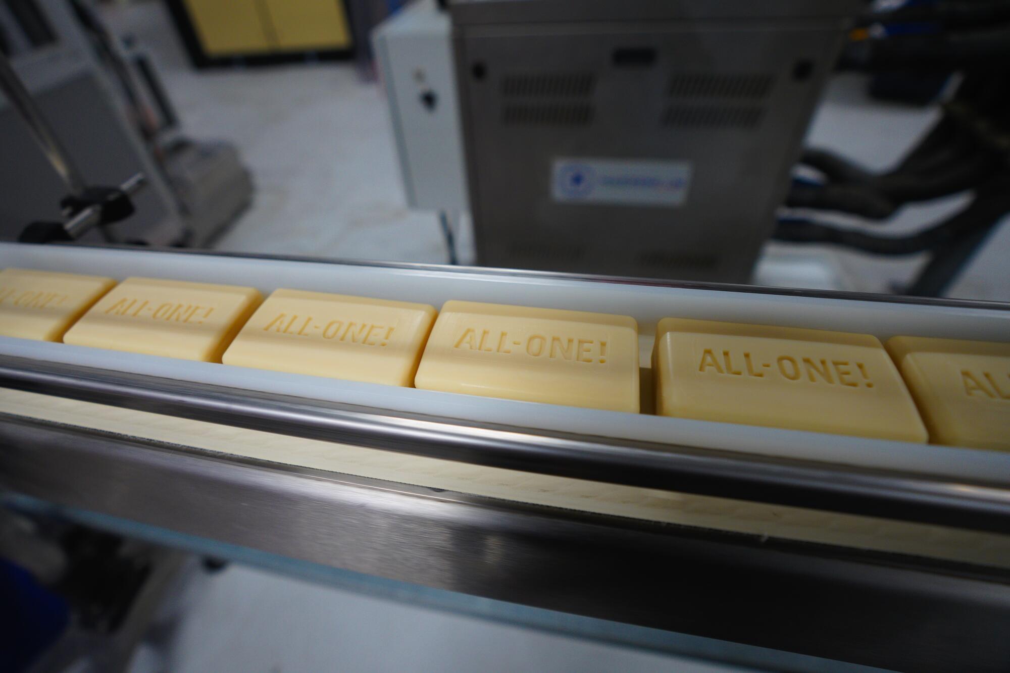 Several cream-colored bars of soap stamped "All-One!" traveling along a factory production line