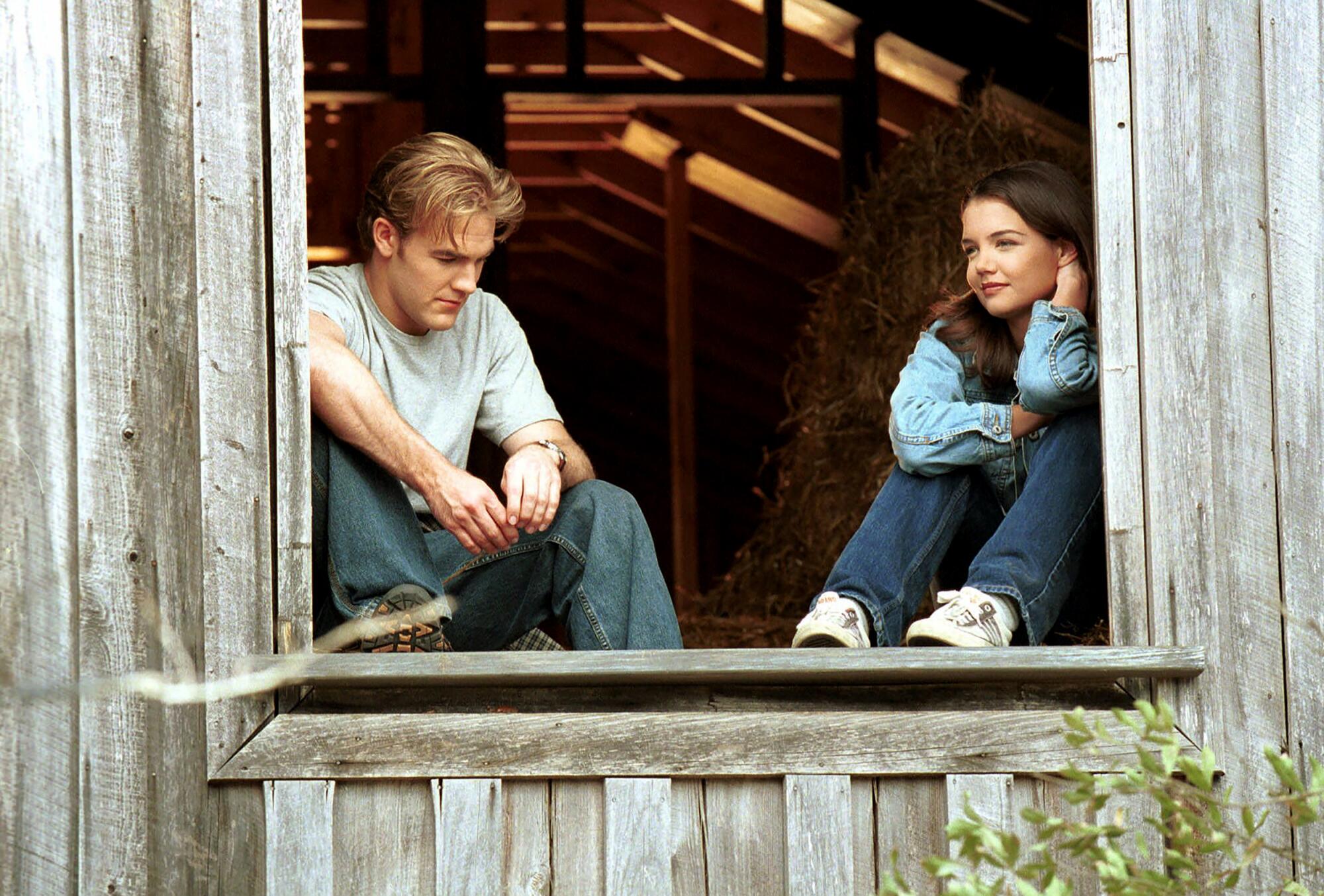 A blond teenage boy and a dark-haired teenage girl sit in the opening of a weather-worn wooden barn.