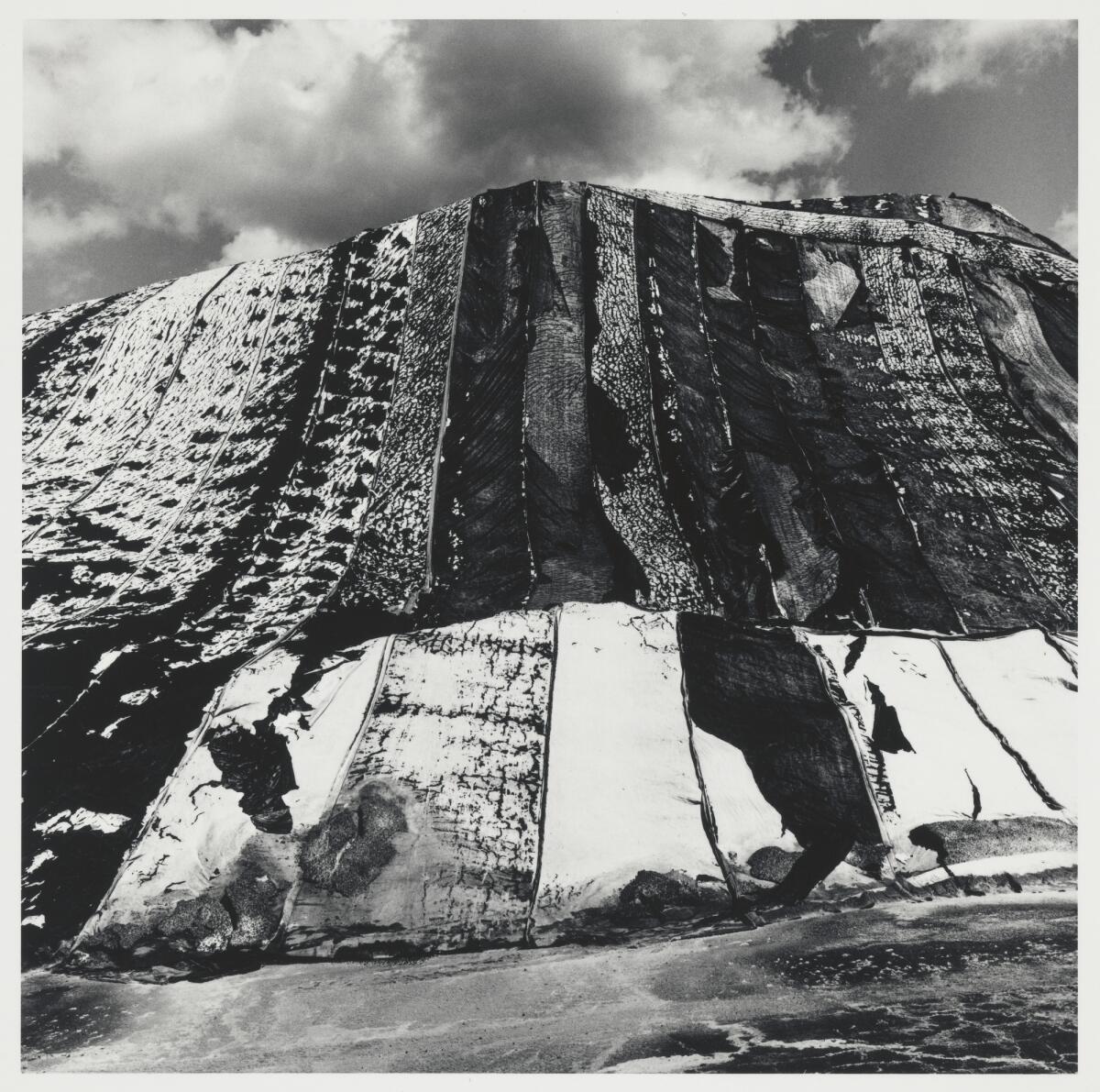 A black-and-white photograph of a salt pile with a sky and clouds in the background.