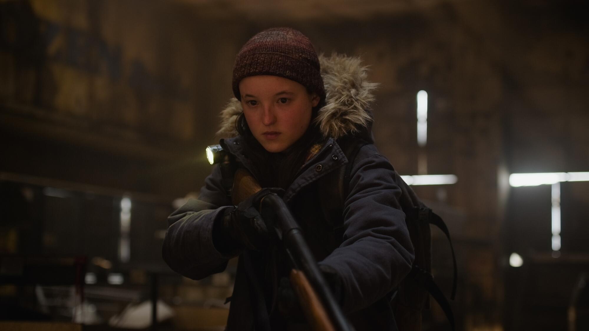 Bella Ramsey in a beanie and coat holding a shotgun.