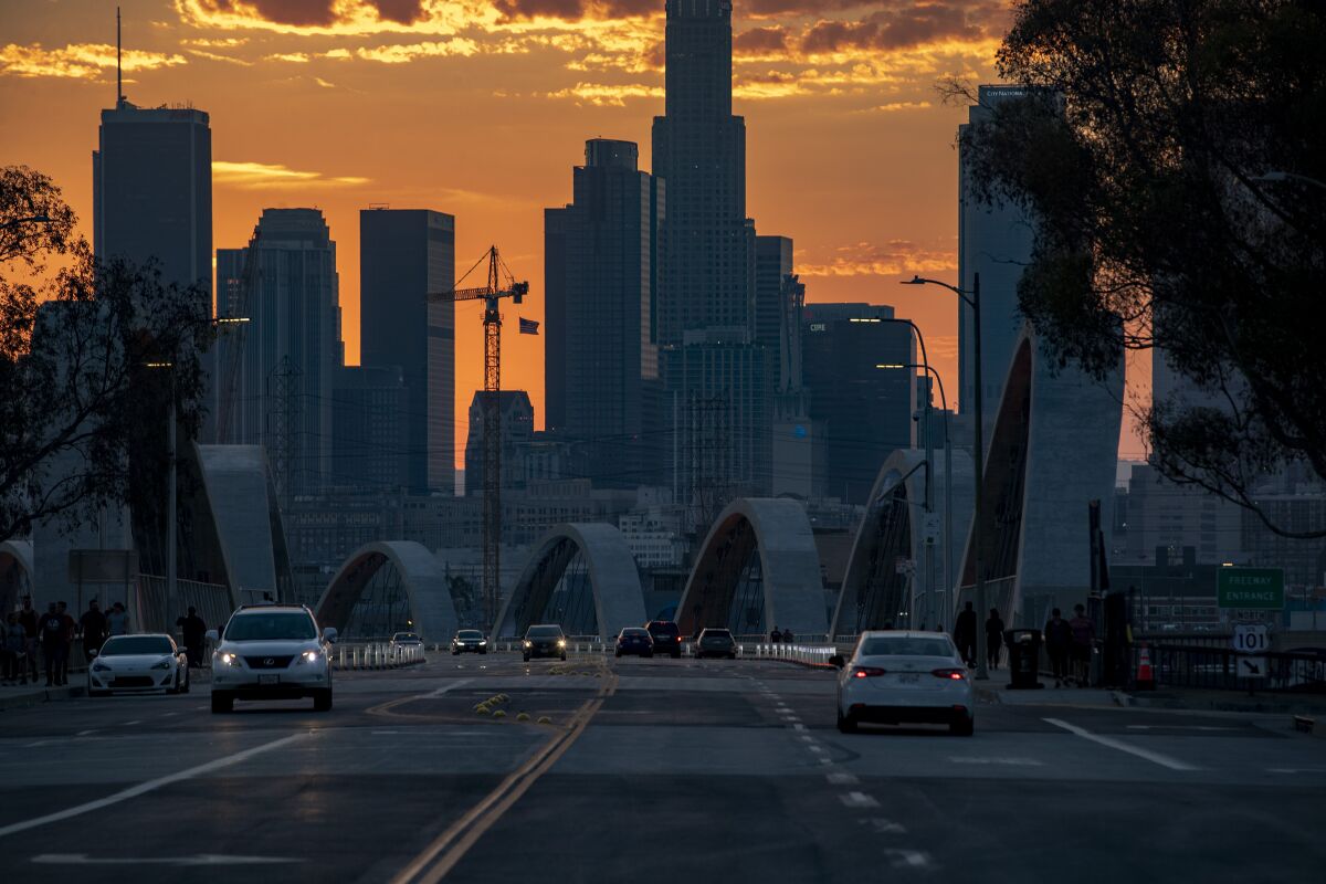 Clouds float in a peach colored sky behind city buildings at sunset. In the foreground cars drive over a bridge.