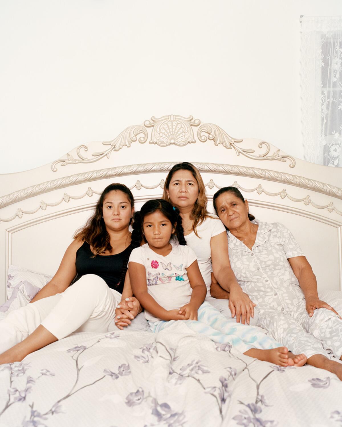 A family of four Latina women is seen sitting on an ornate white bed.