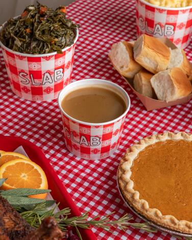 Collard greens, gravy, pumpkin pie and dinner rolls on a red and white checked tablecloth.