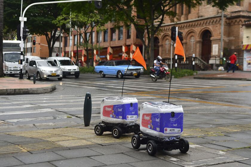 Two delivery robots, looking like little coolers on wheels with a flag attached, roll down a Medellin street