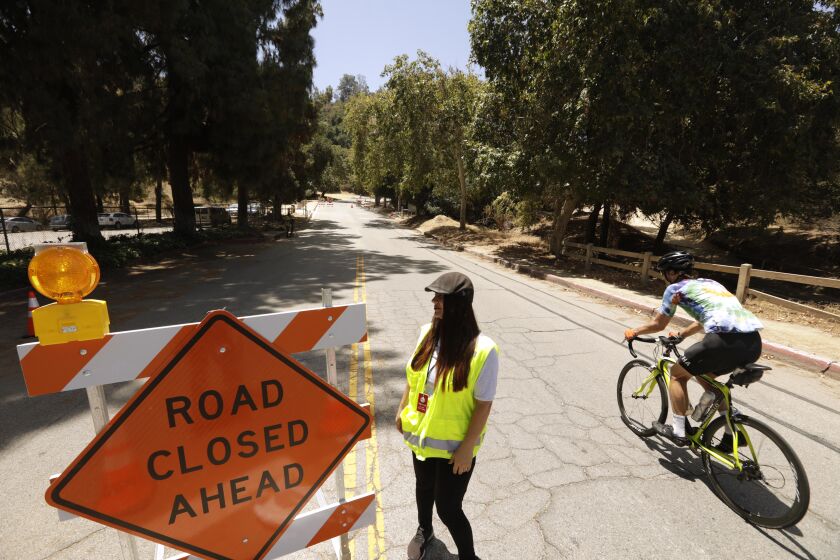LOS ANGELES, CA - JUNE 29, 2022 - - A bicyclist rides past city worker Emma Delgado to head down a one-third mile portion of Griffith Park Drive that is indefinitely closed to car traffic to improve safety for pedestrians and cyclists in Griffith Park in Los Angeles on June 29, 2022. Delgado was on site to tell motorists that they could not go down this portion of Griffith Park Drive. The road is known to draw high-speed drivers who are looking for a shortcut, avoiding the 134 and 5 freeways. The temporary closure has been in place since Monday, June 27 and will carry on indefinitely until more permanent changes to the park's infrastructure are in place. (Genaro Molina / Los Angeles Times)