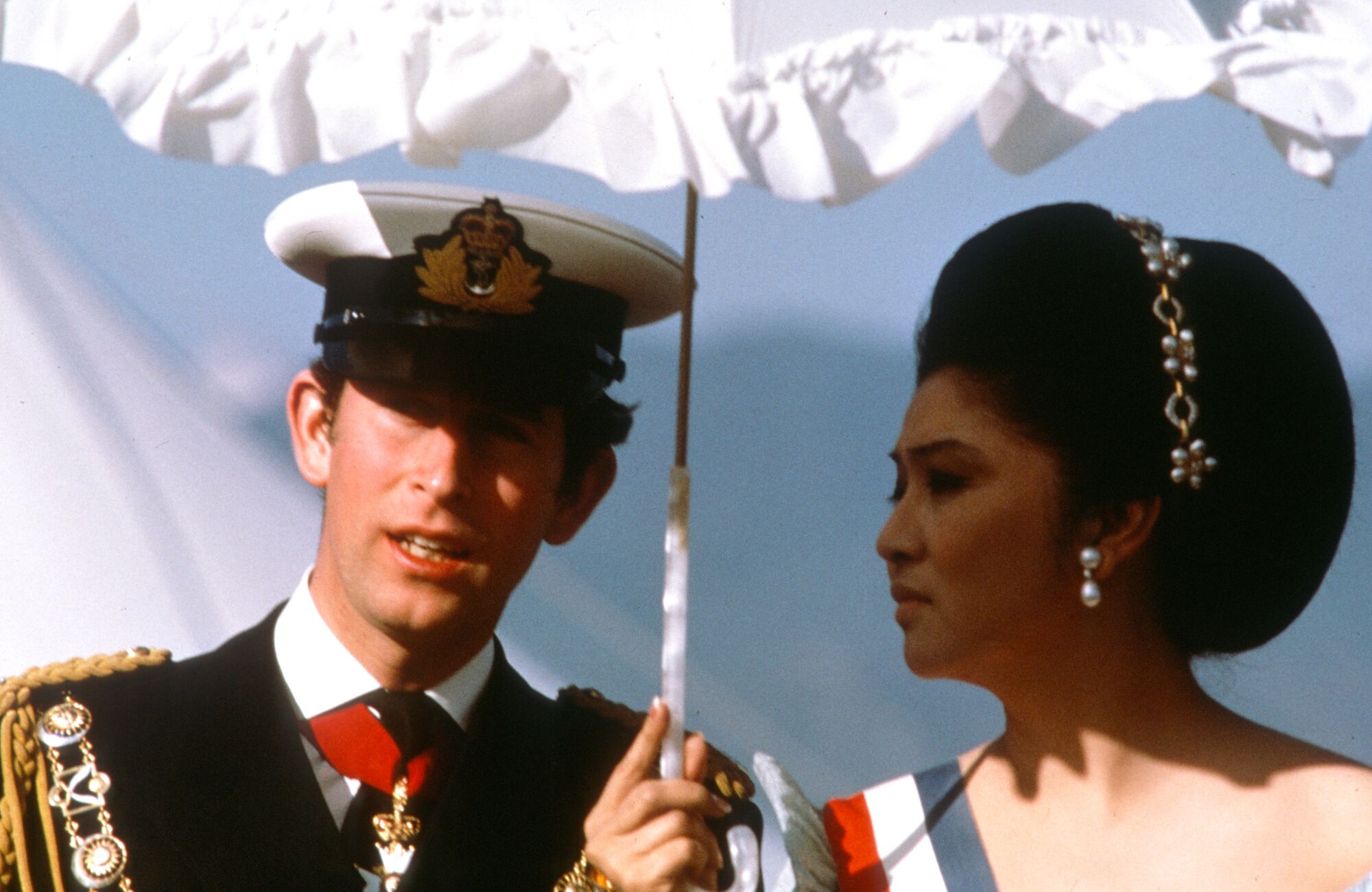 A man in military uniform and hat, left, next to a woman wearing pearl earrings and hairband and holding a white parasol 