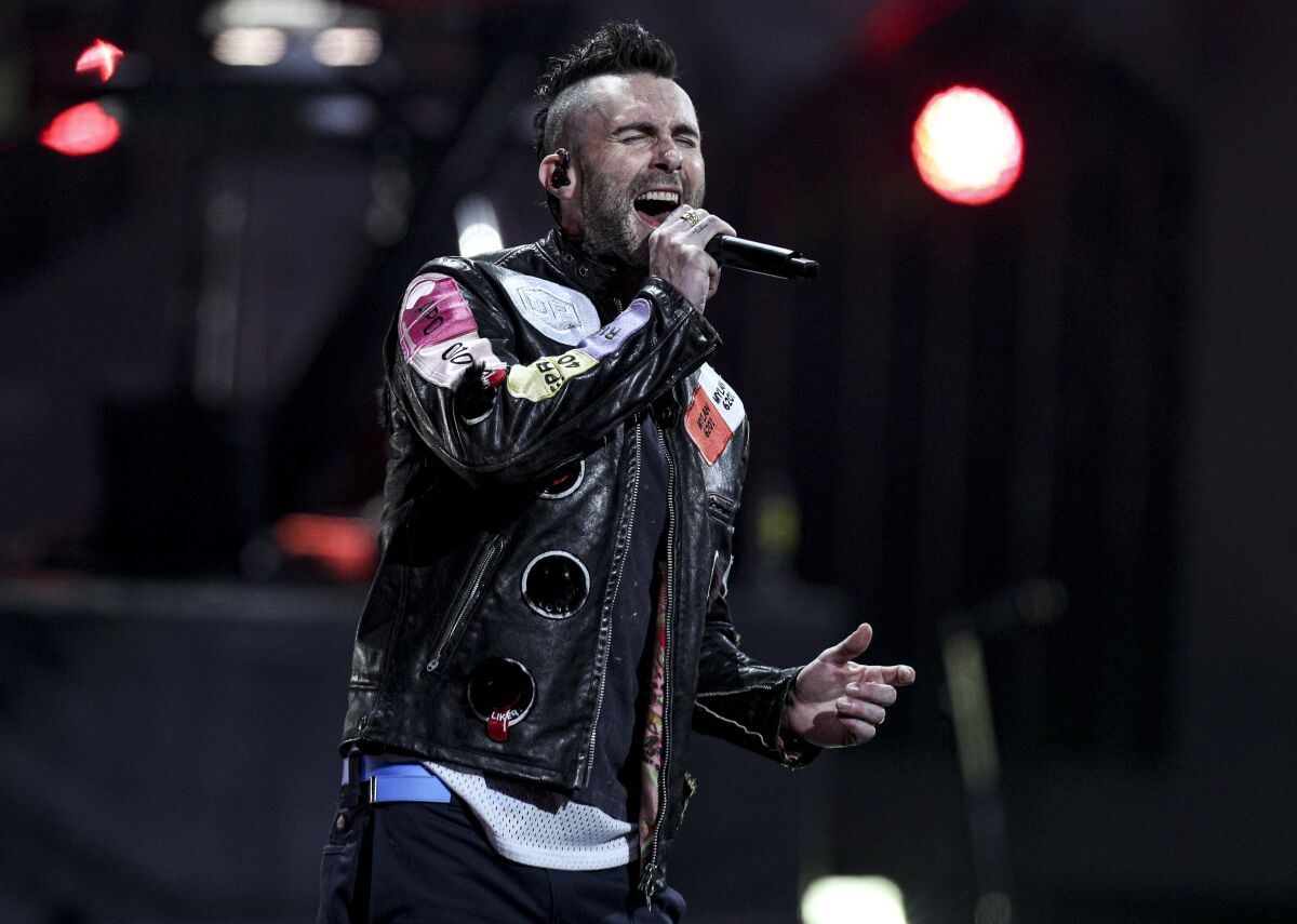 Adam Levine wearing a leather jacket with patches and singing into a microphone on stage.