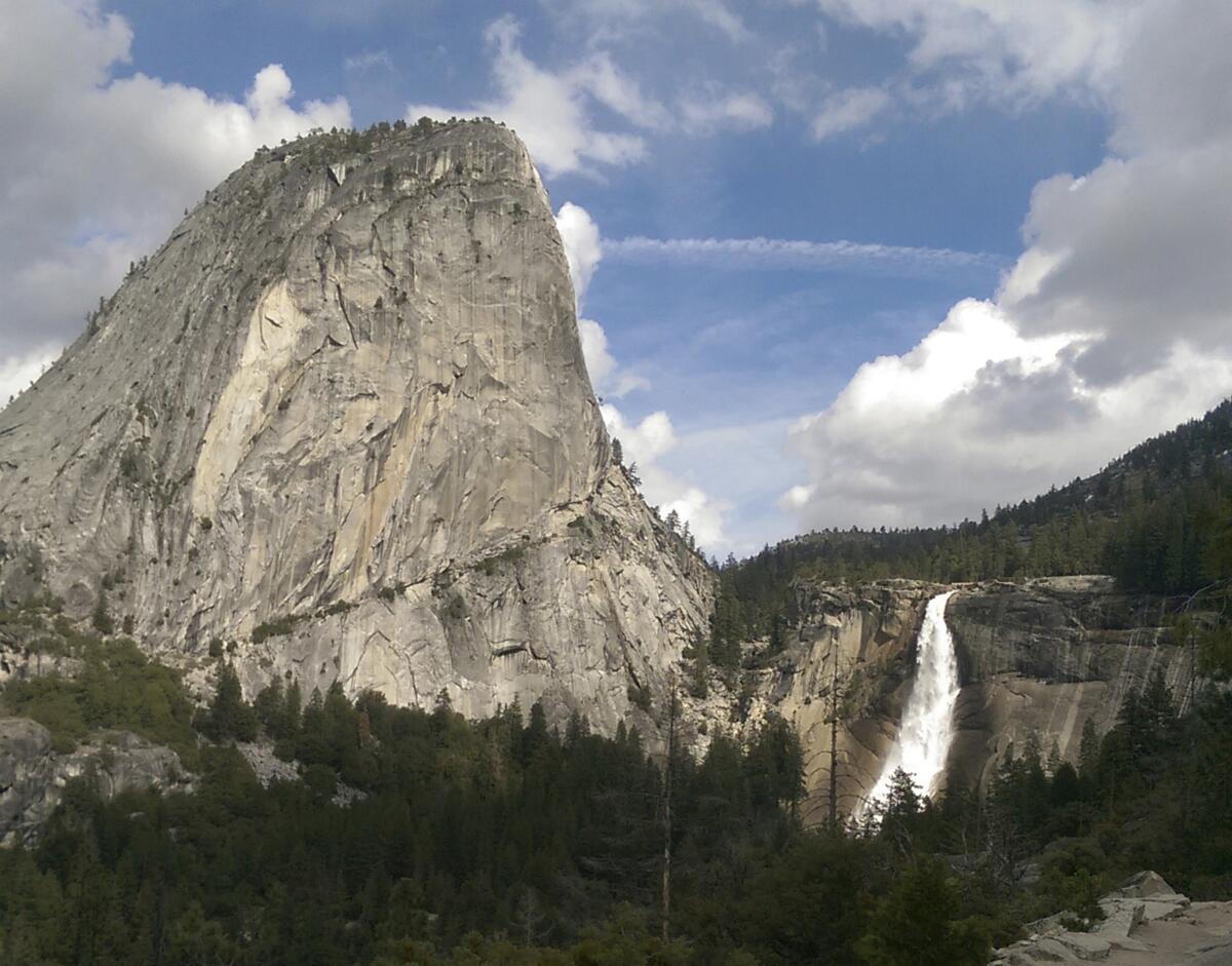 A waterfall flows next next to a mountain with evergreen trees in the foreground.