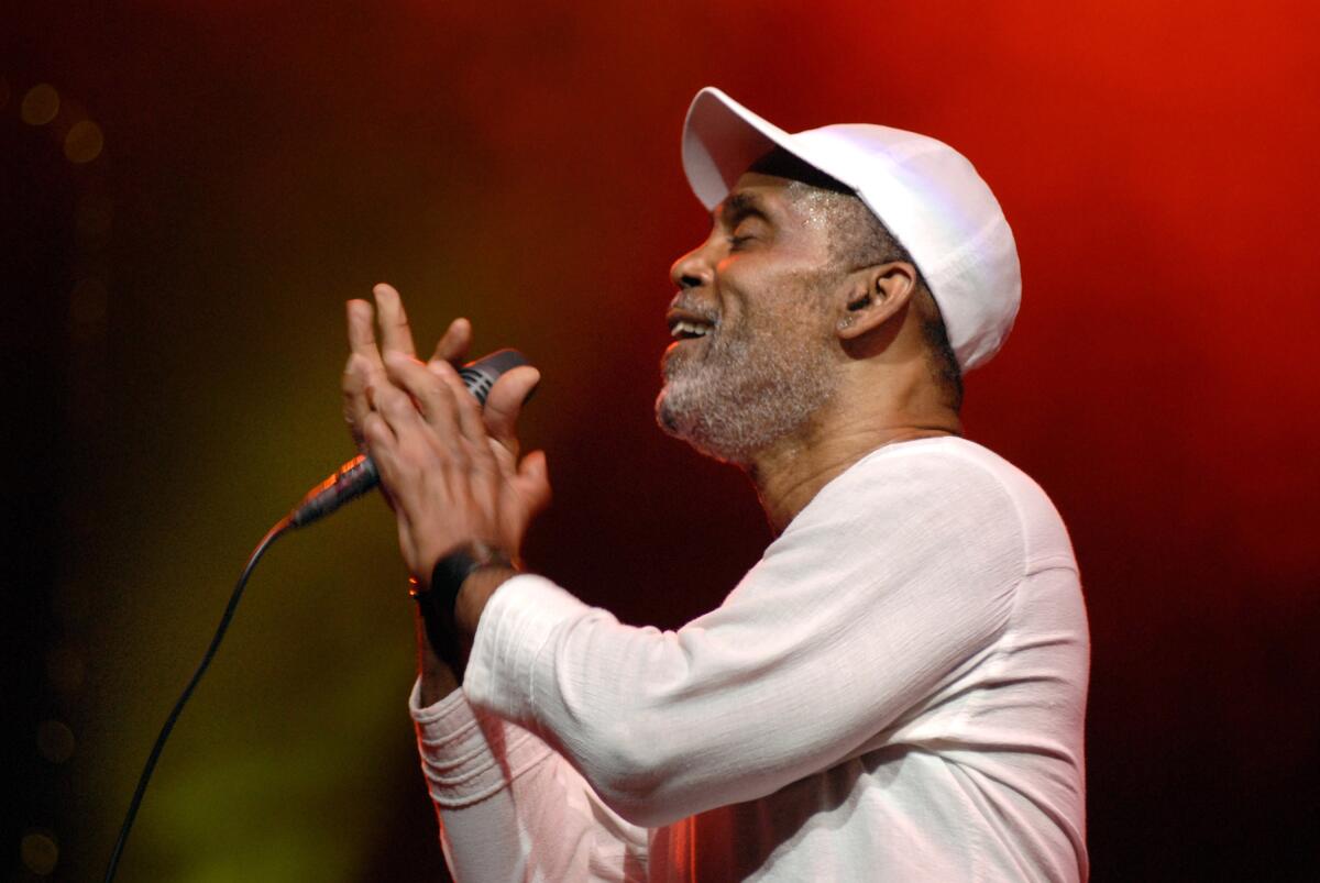 Frankie Beverly holds a microphone while wearing a white cap and shirt 