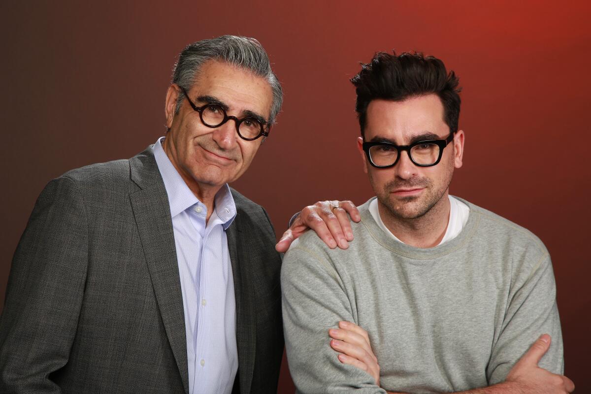 Eugene Levy, wearing a sports coat, shirt and glasses, standing next to Dan Levy, in a grey sweater and glasses.