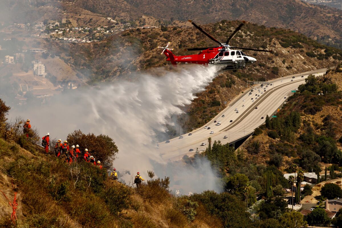 A helicopter drops water on a steep slope burning above a freeway