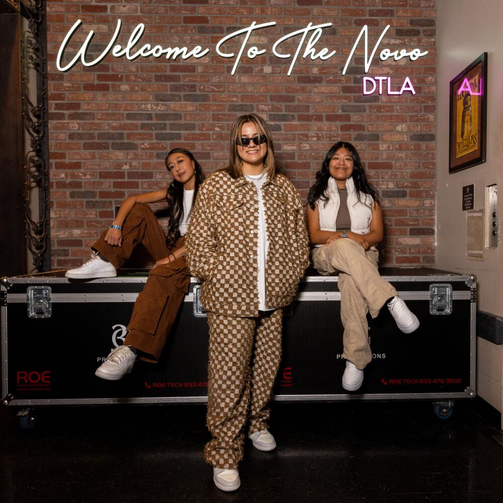 Two women sit on a trunk while another woman stands in front of them below the words Welcome to the Novo DTLA