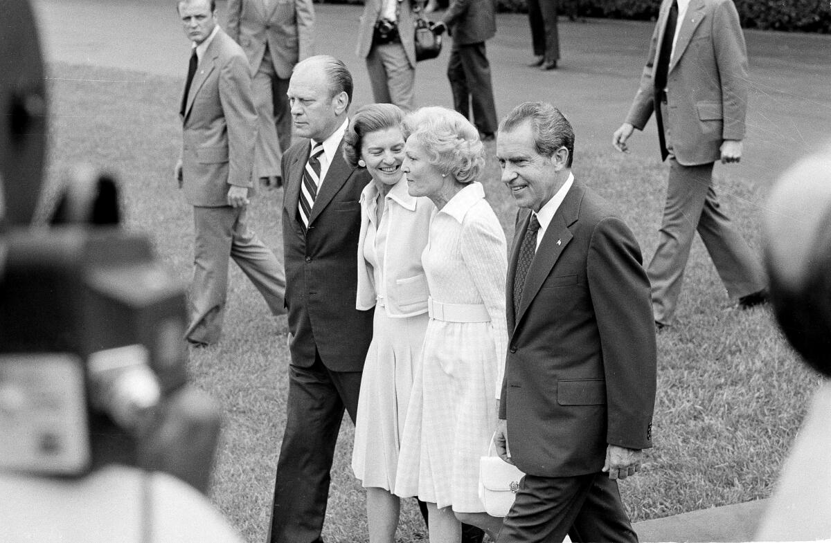 A black-and-white photo of Gerald and Betty Ford walking side-by-side with Richard and Pat Nixon, as security and media watch
