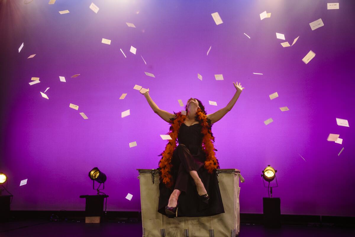 A woman in a fur stole sits onstage with her arms extended to the sky as pieces of paper flit from the ceiling