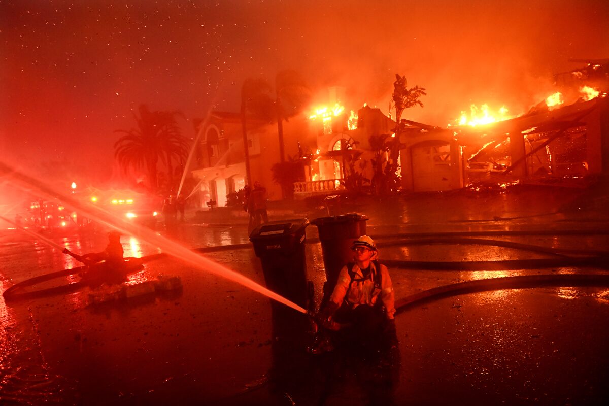 Firefighters spray water from hoses on a street with homes engulfed in flames and a red glow