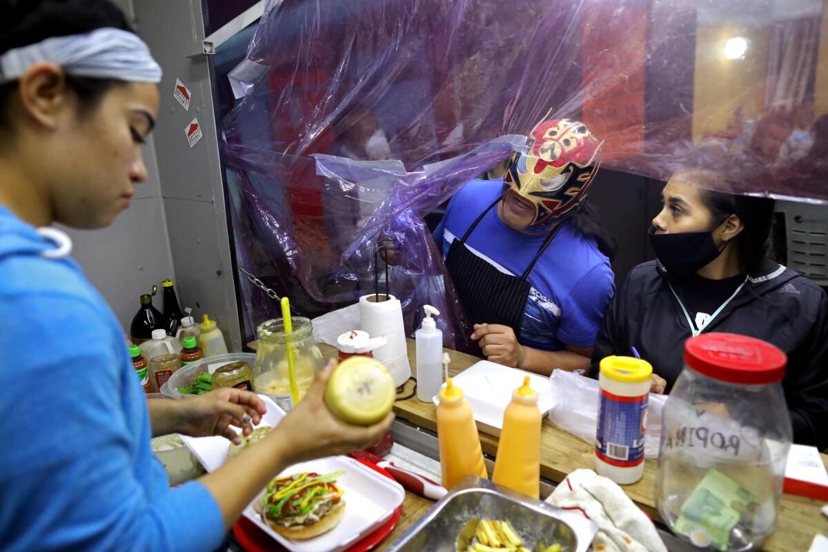 A luchador peaks his head inside a food track as a woman stands behind the grill.