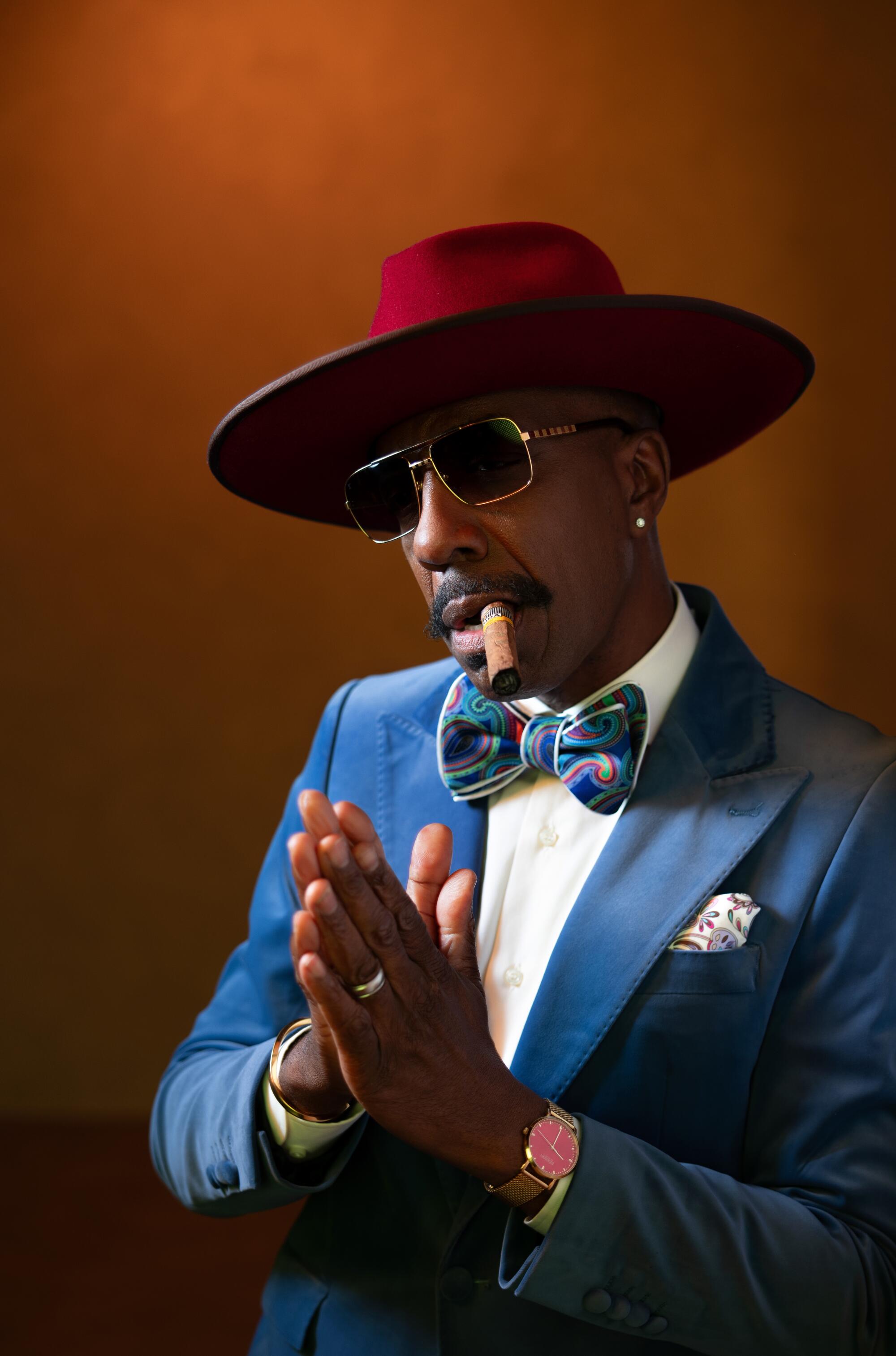 JB Smoove poses for a portrait in a wide-brimmed hat and sunglasses, with a cigar in his mouth.