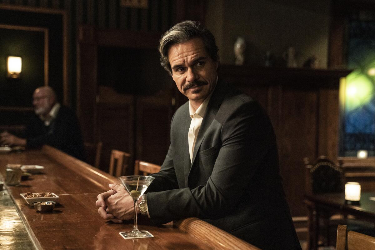 A mustachioed man in a suit sits at a bar with a martini