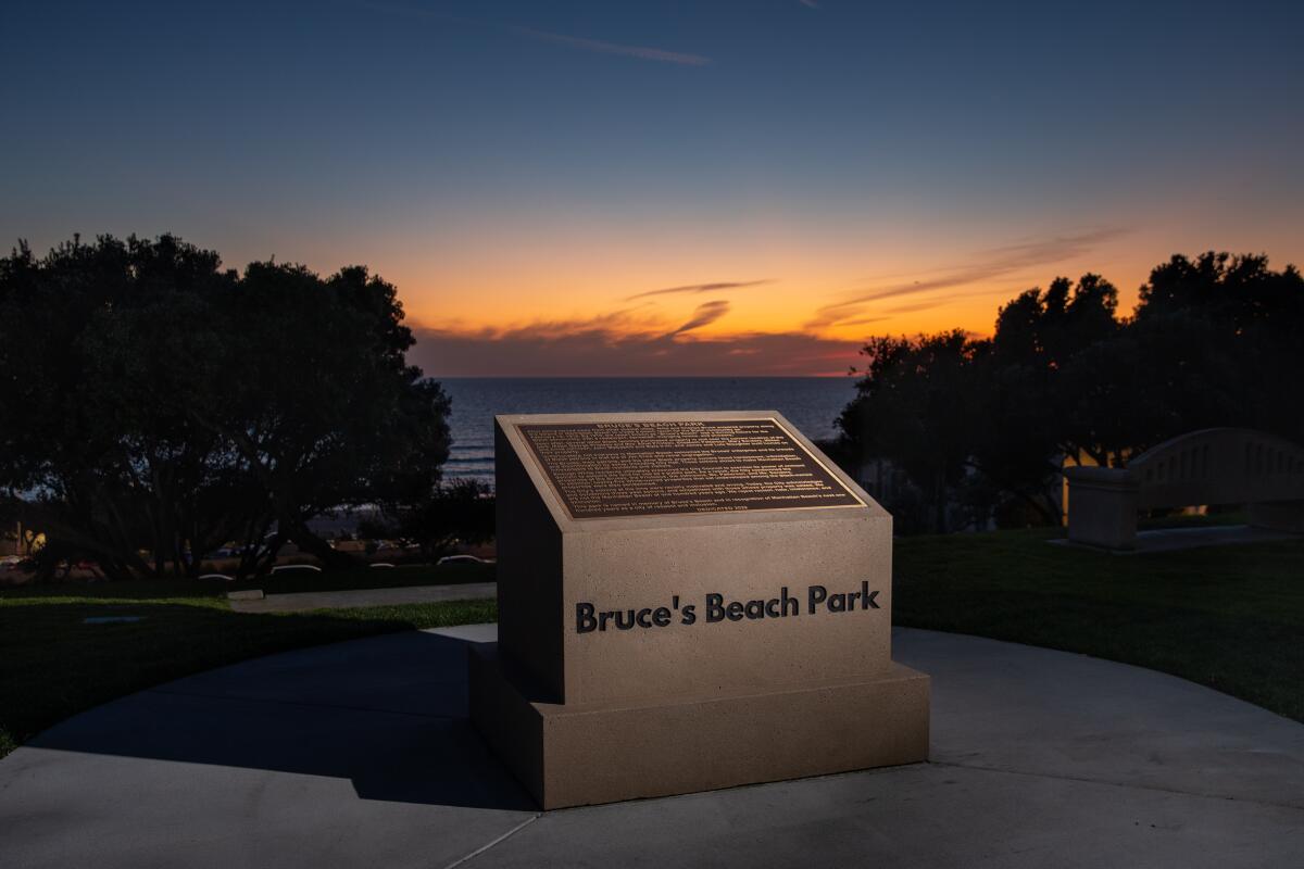A monument with a plaque is shown at sunset with a beach in the background.