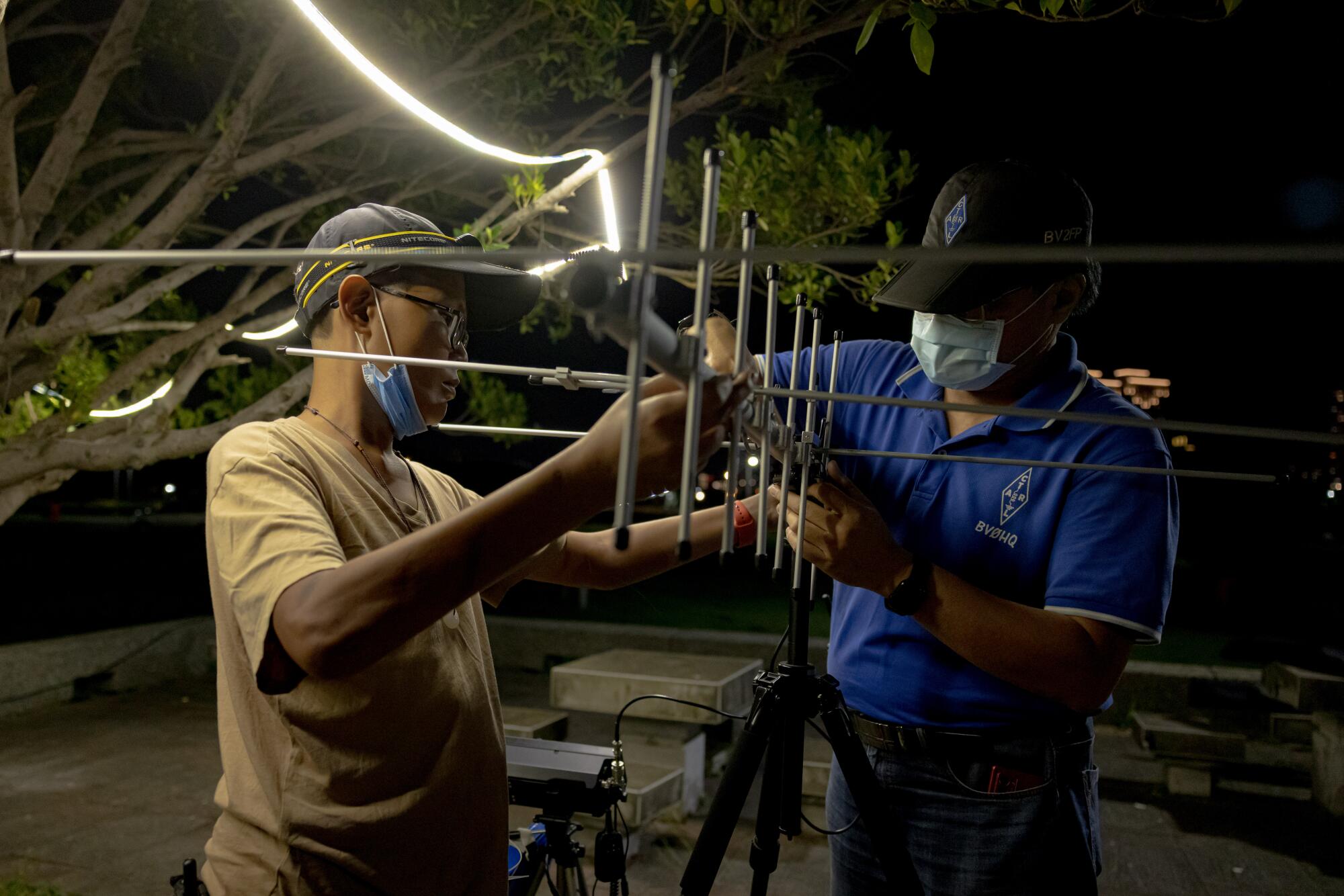 Two men hold an antenna near a tree strung with lights 