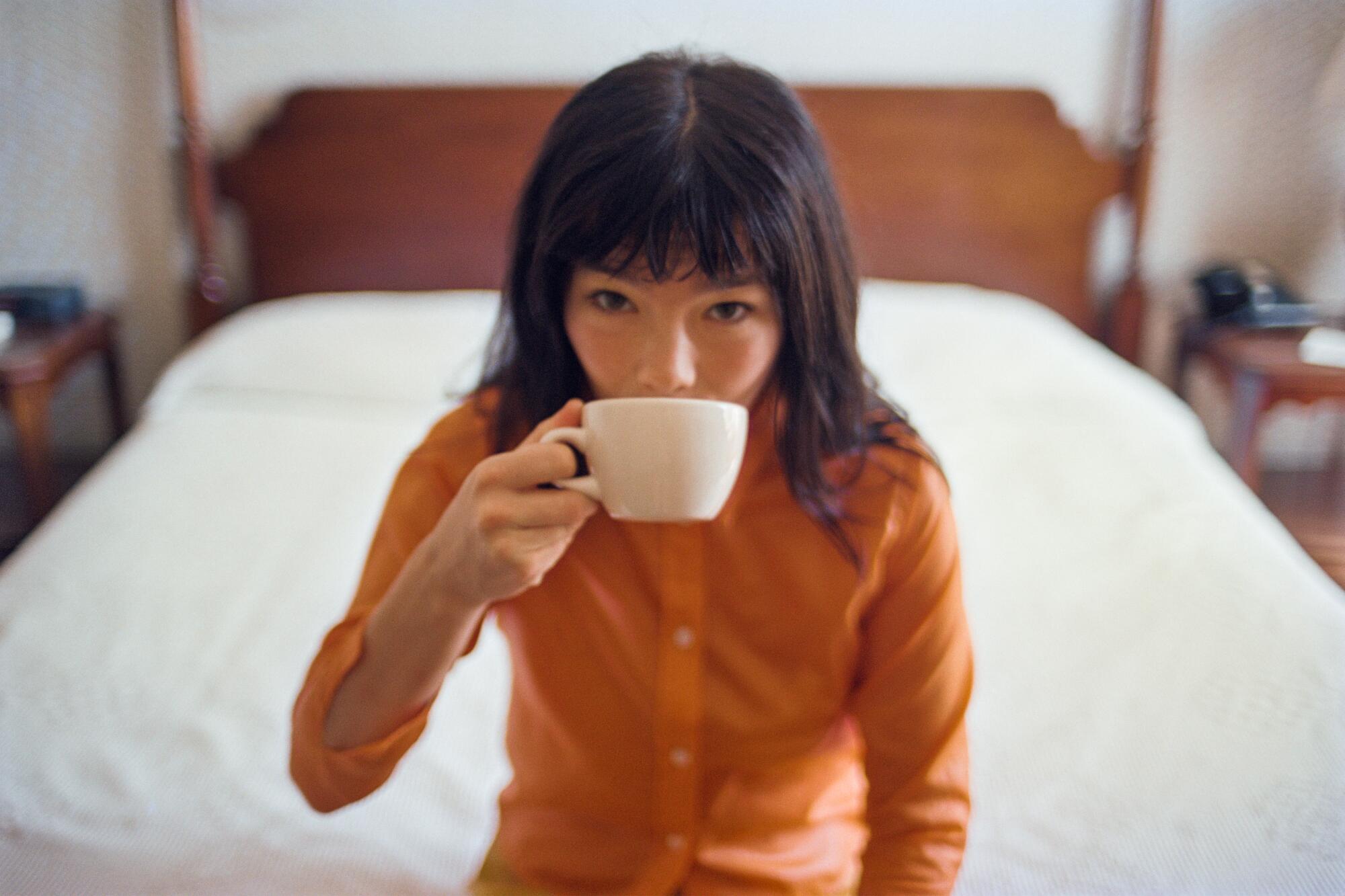 Björk sits on the edge of a bed, wearing an orange sweater and drinking from a white coffee cup.