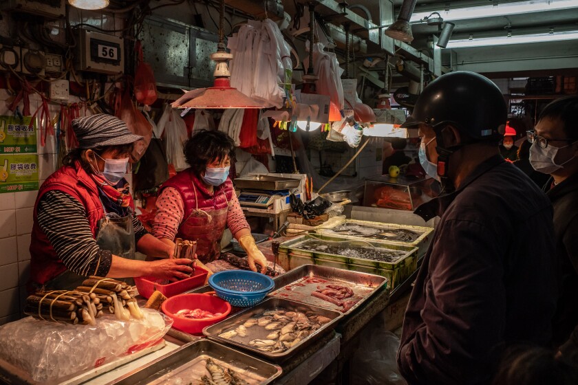 Two people in masks handle seafood that fills trays atop a table. Two others peruse the goods.