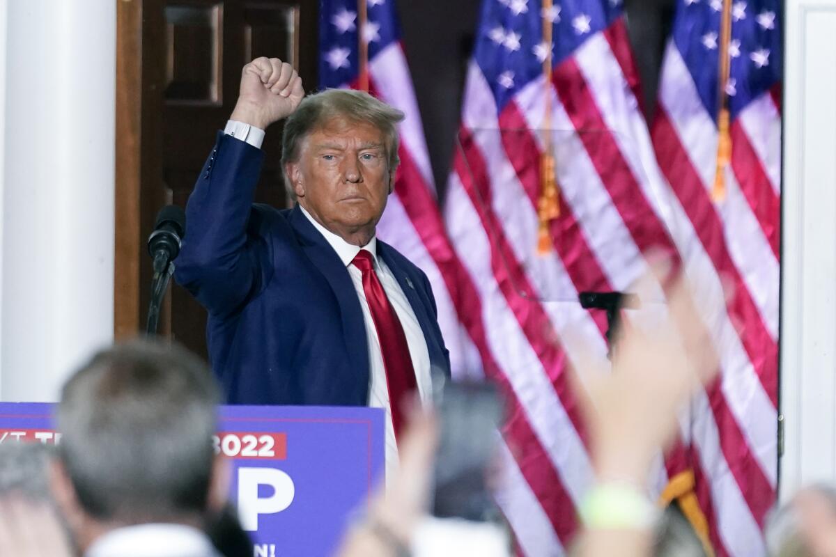 Former President Trump raising a fist as he stands in front of multiple American flags and faces a crowd