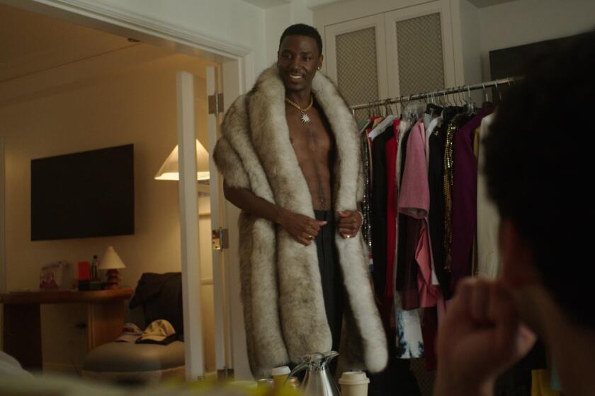 A shirtless man in a fur coat standing before a rack of clothes.