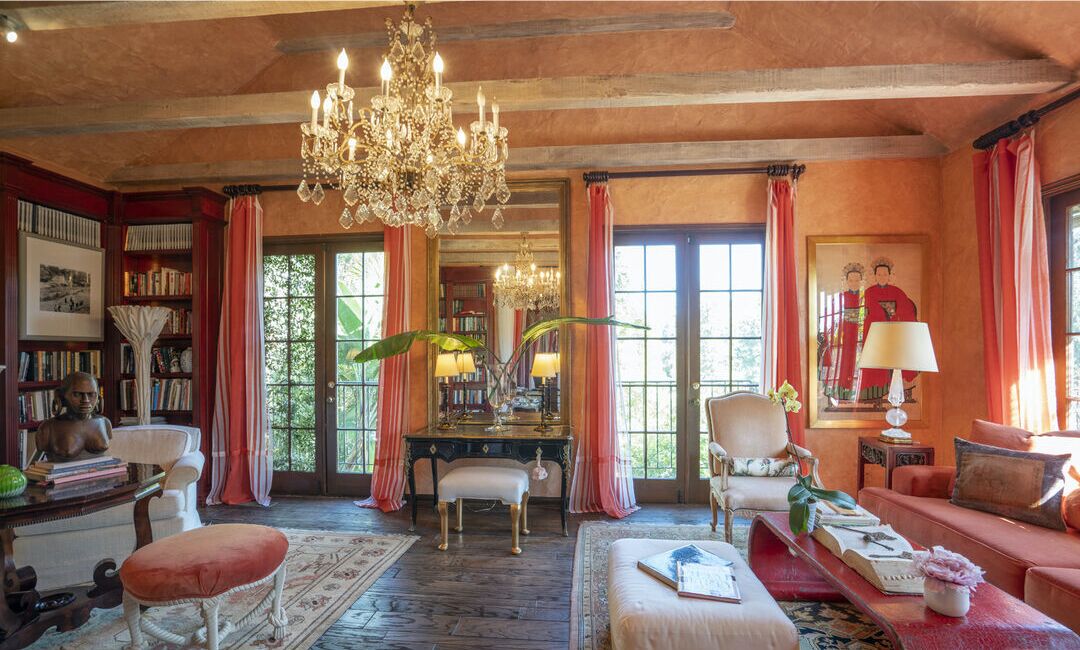 The lounge is in orange shades with a chandelier, bookshelves, furniture and windows overlooking foliage.