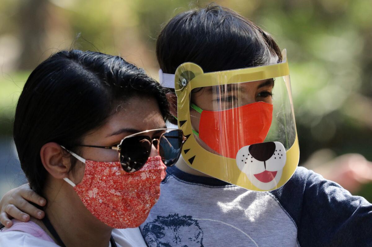 A woman wearing a red mask and sunglasses carries her son, who has a plastic tiger face shield over his mask