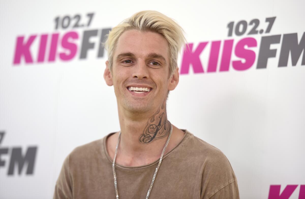A man with blond hair and a neck tattoo smiles while standing in front of a backdrop