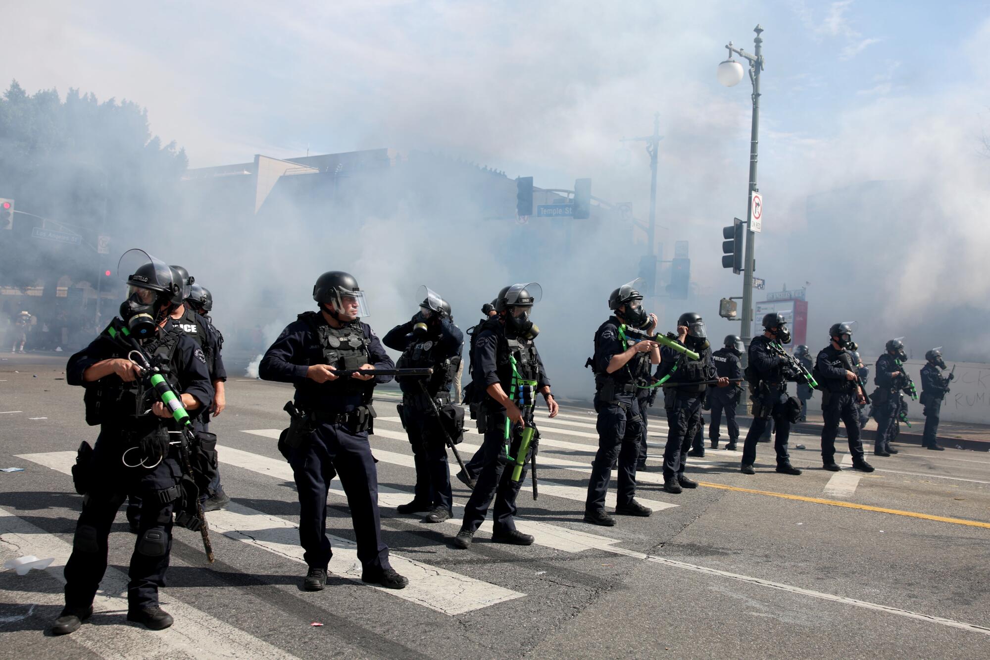 A row of police officers wearing black stand in a crosswalk with smoke billowing behind them