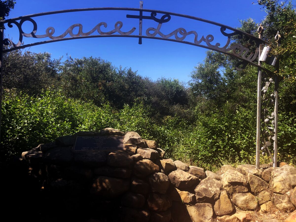 A curving metal sign above rocks on a tree-covered hillside reads "Peace, Love, Joy."