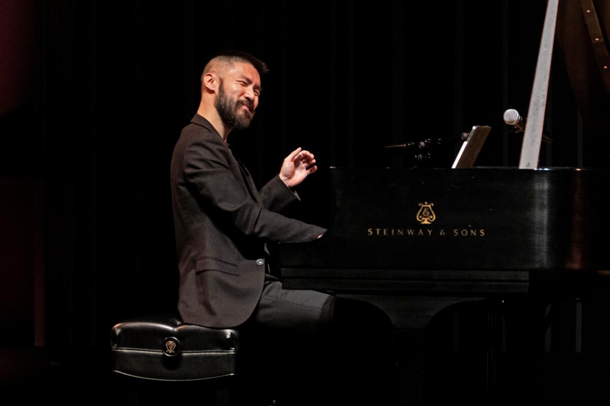 Conrad Tao, in a black suit, is seen playing a black grand piano on stage.