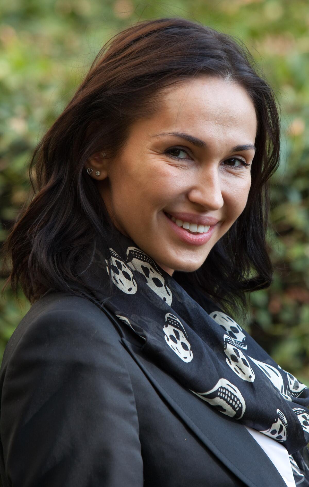 A dark-haired woman in a jacket and scarf emblazoned with skulls smiles with greenery in the background
