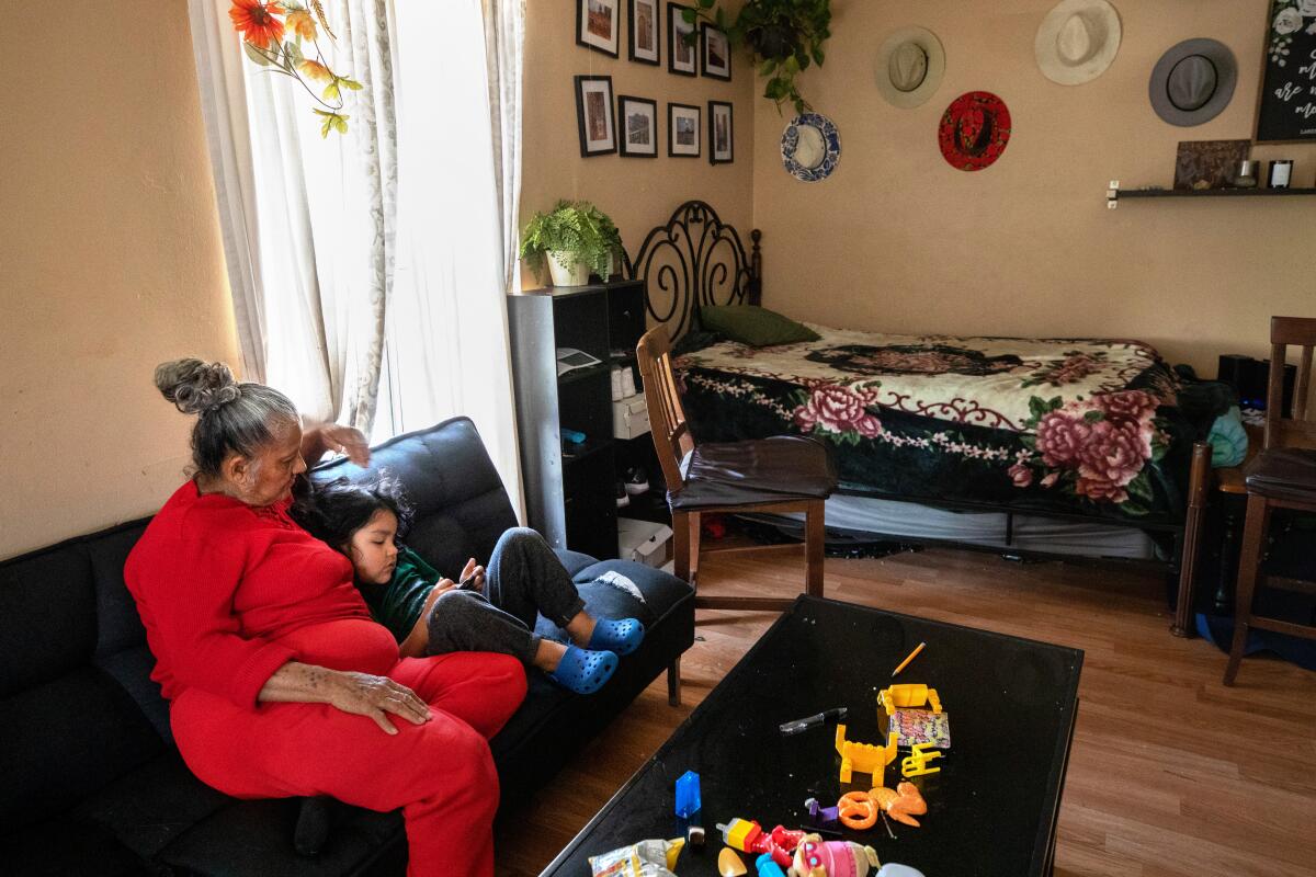 A woman sits on a dark sofa with a young child curled up against her.  A bed and other furniture line a wall behind