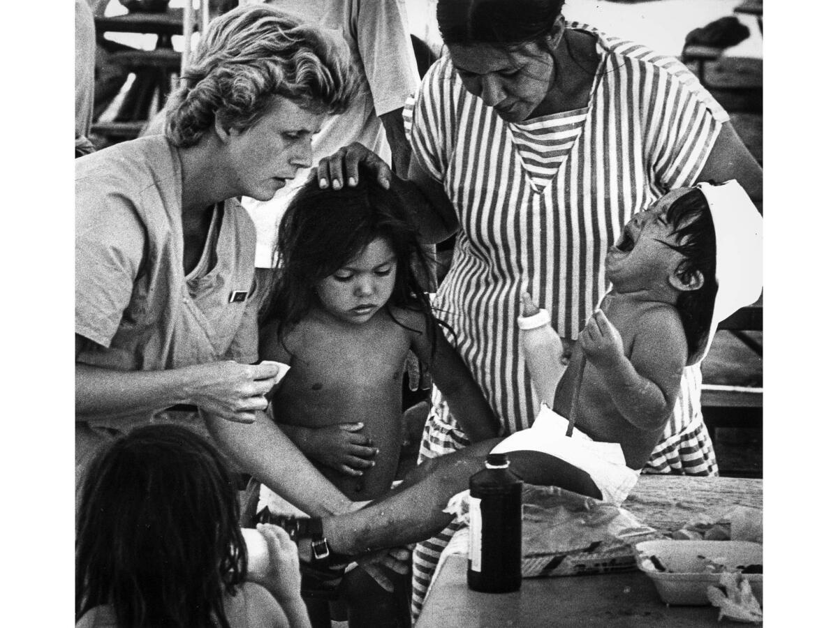 July 15, 1987: Volunteer Janice Estes, a medical assistant from Fullerton, examines a 2-year-old boy with sores on his legs. The boy's mother and sister look on.