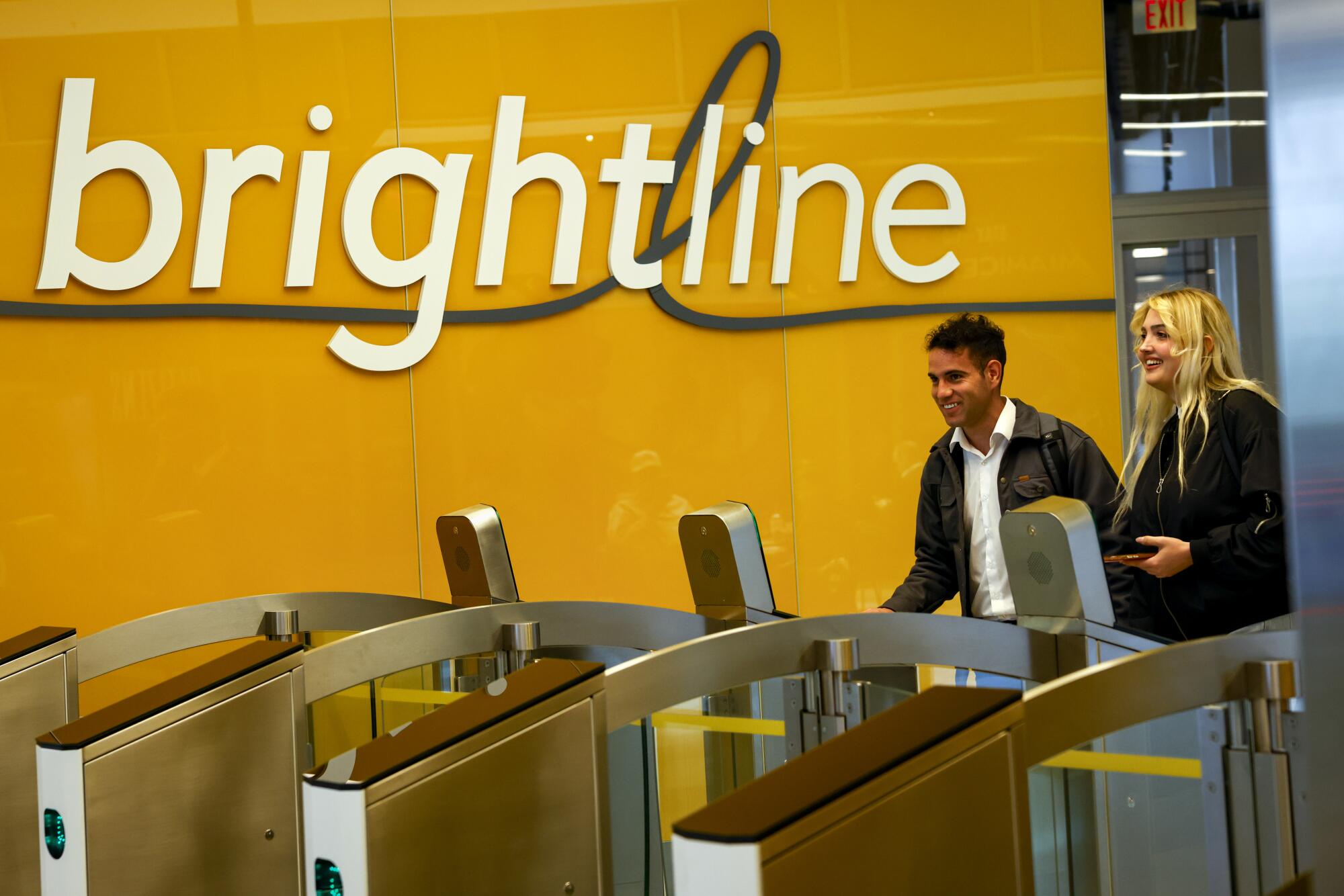 A man and a woman approach electronic gates, with a sign saying "brightline" on a yellow wall 