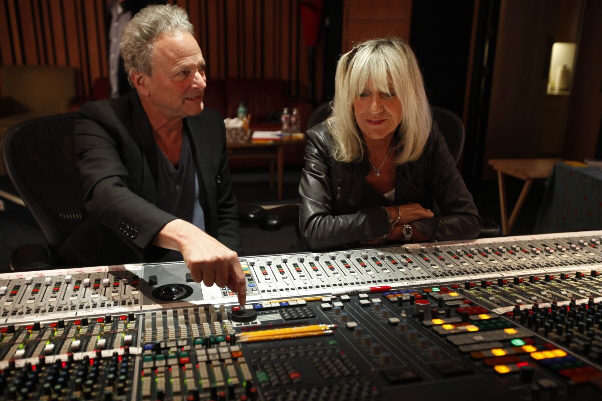 Man in black blazer and woman in black leather jacket sit in front of music studio sound mixing board. 